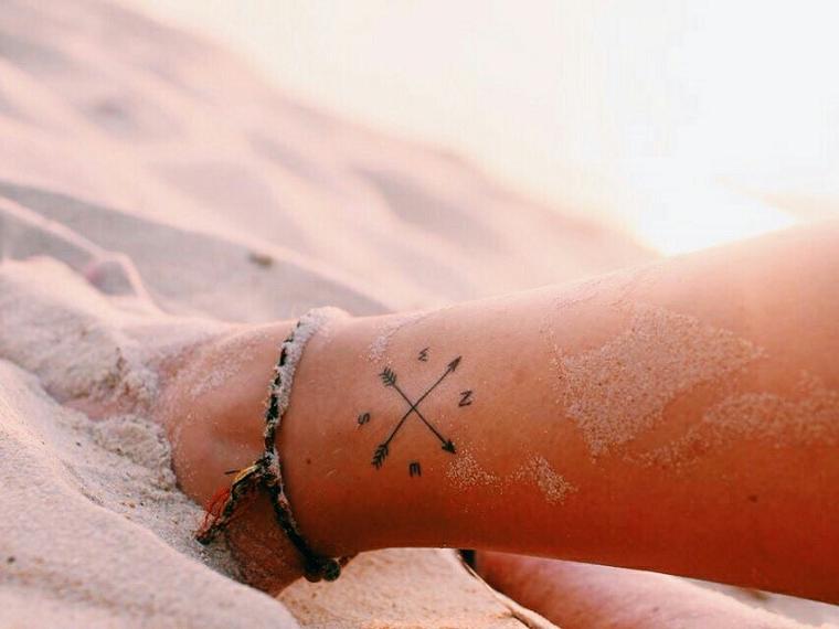 Tattoo caviglia, disegno tatuaggio bussola, donna con braccialetto sulla caviglia, piede immerso nella sabbia