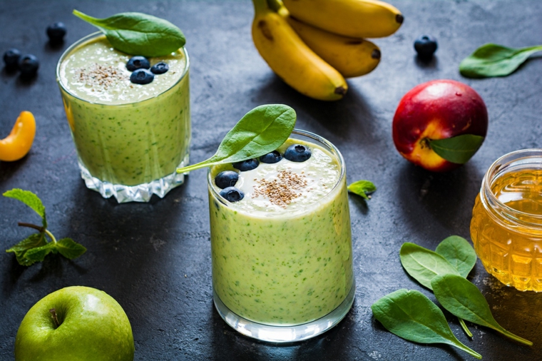 Bicchiere di vetro con smoothies verde, ingredienti sparsi su un tavolo, pesche e baby banane 