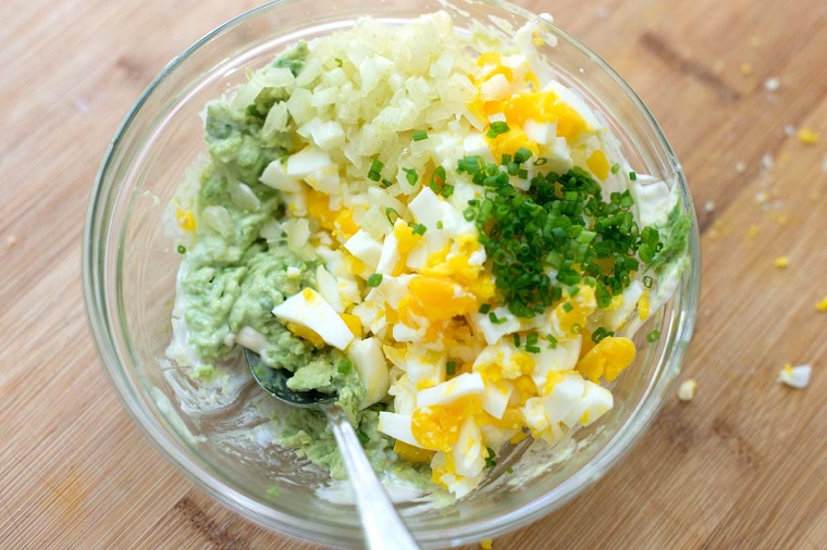 Avocado come si mangia, ciotola di vetro con uova cotte e pezzettini erba cipollina 