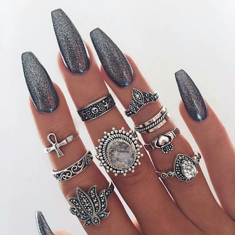 Manicure unghie a stiletto di colore argento glitter, tanti anelli di metallo sulle dita