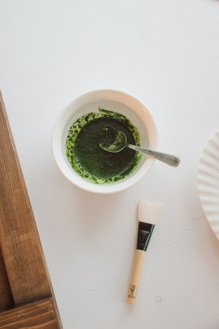 Ciotola con maschera al tè verde matcha, pennello su un tavolo di colore bianco